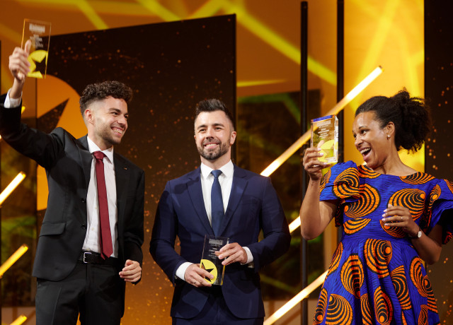 Nela Riehl, Member of the European Parliament for Volt, smiles joyfully while holding up her MEP Award on stage. She is wearing a vibrant blue and orange patterned dress. Two men in suits stand beside her, also holding awards and smiling, with a celebratory atmosphere and bright stage lighting in the background.