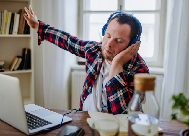 Young man with down syndrome sitting at a desk at home, using a laptop and listening to music with headphones
