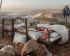 Source: https://mondoweiss.net/2024/11/photo-essay-the-silent-displacement-in-the-west-bank/#:~:text=As%20the%20world%20has%20focused,Bank%20following%20October%207%2C%202023. A photograph of two metal bed frames placed outdoors on a dry, rocky hillside. In the foreground bed, a young child is asleep under a thick, patterned blanket and pillows. The surrounding landscape is arid and mountainous, with a light haze or dust hanging in the valley behind them. Various items, including a water bottle and a jacket hanging from a wooden post, suggest a makeshift living space. The lighting is soft, indicating the early morning or late evening.